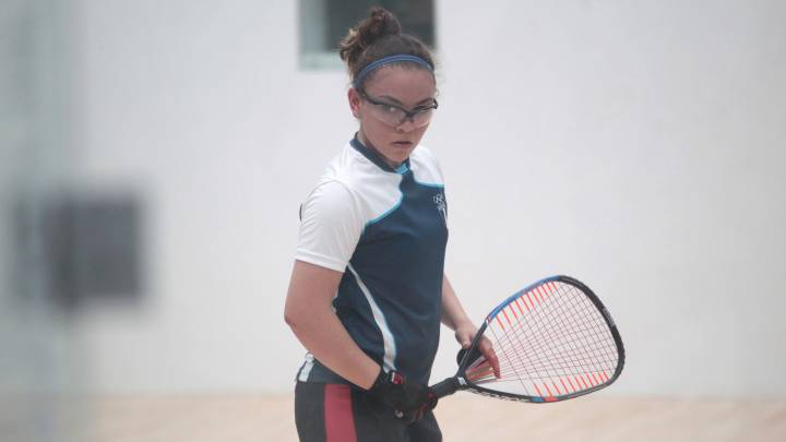 Mujer raquetbolista, viste un short gris y playera blanca con gris, tiene una raqueta en la mano.