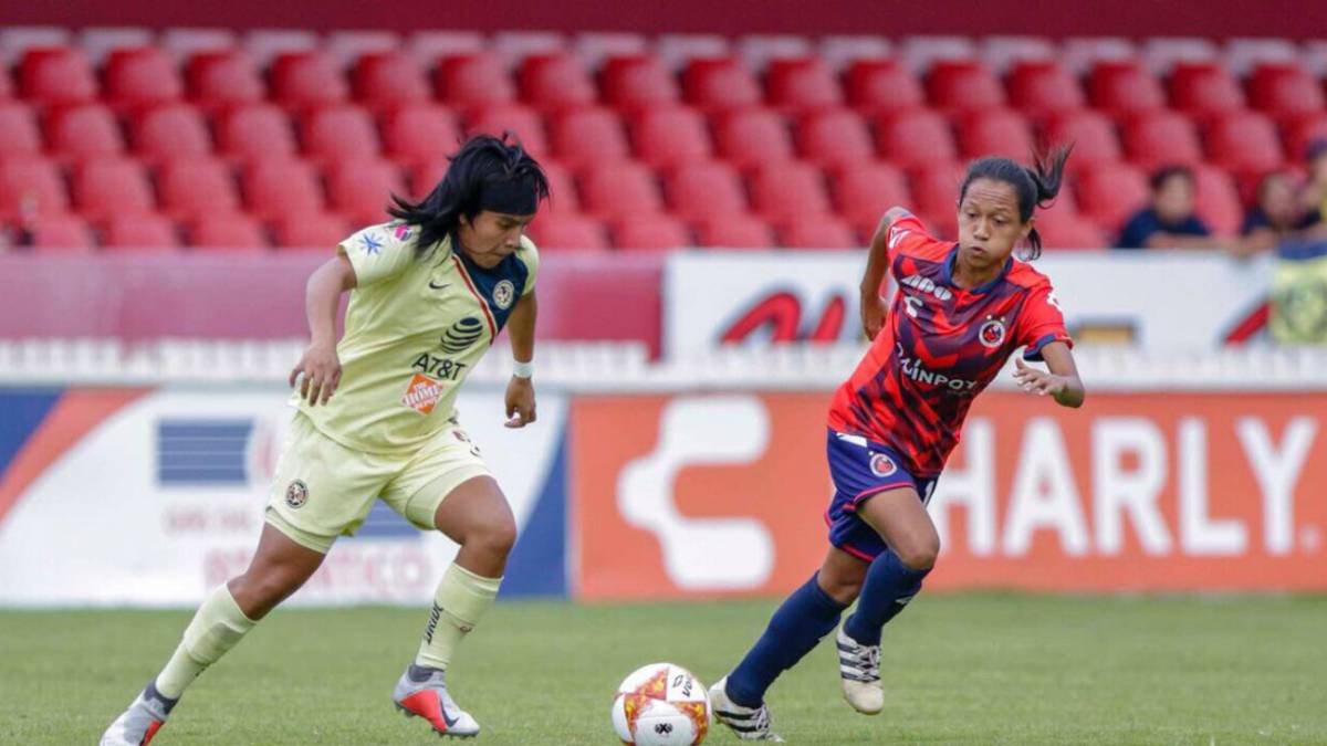 Jugadoras de fútbol disputando el balón, una está vestida de amarillo y la otra tiene short azul con playera en rojo y azul
