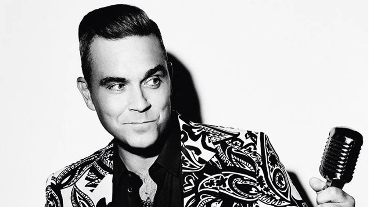 El cantante británico Robbie Williams en una imagen en blanco y negro en la que aparece con una media sonrisa mirando un micrófono que tiene en la mano y con un llamativo traje. Foto Instagram @robbiewilliams