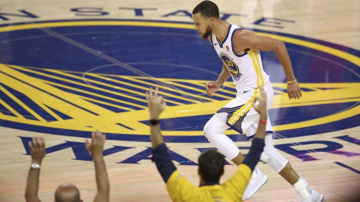 Stephen Curry corre hacia su canasta mientras dos aficionados levantan las manos. El jugador mira al suelo justo a la mitad de la duela donde se encuentra el logo de Golden State