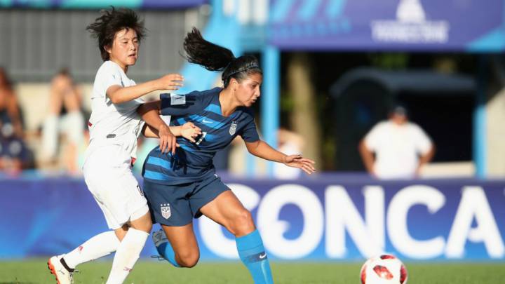 2 jugadoras de futbol disputando el balón, una con uniforme blanco otra con unifome azul