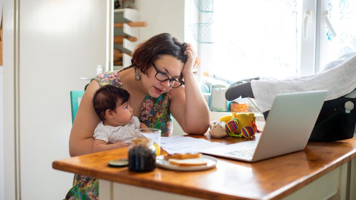 Una mujer cargando su bebé y revisando documentos frente a su computadora