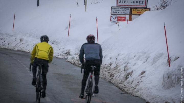 Varios ciclistas realizan la subida a Alpe d'Huez entre paredes de nieve a los lados de la carretera.