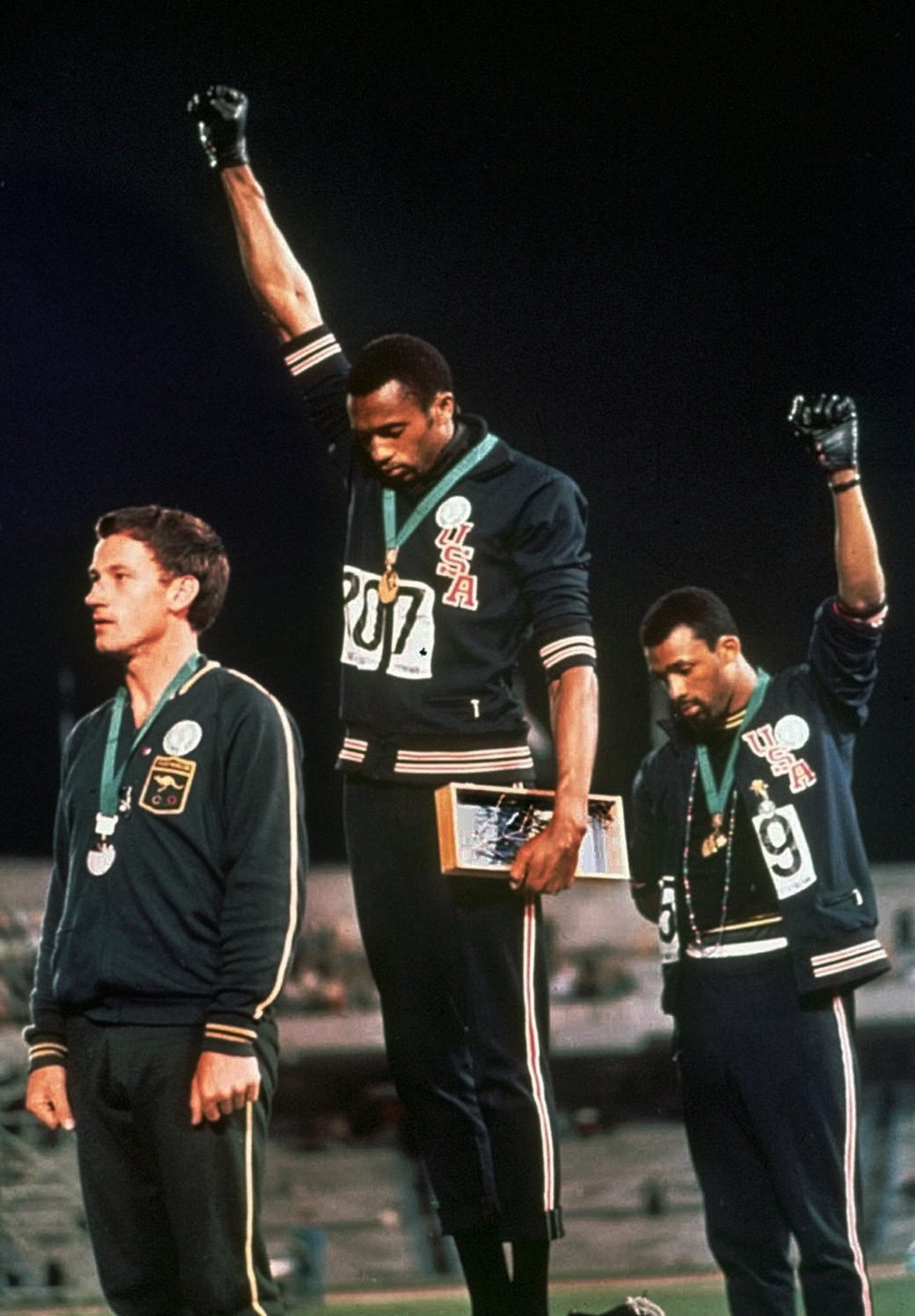 Tommie Smith (centro) y John Carlos (derecha) levantan sus puños con guantes negros en el pódium